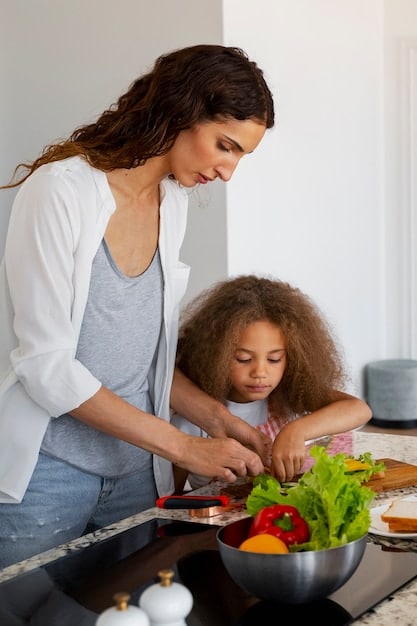 A child happily stirring a bowl of salad ingredients under the supervision of an adult, both smiling and engaged in the cooking process in a well-equipped kitchen.