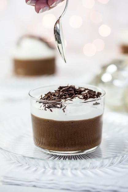 A close-up shot of a creamy chocolate mousse in a clear glass, topped with fresh raspberries and a dusting of cocoa powder. The background features a softly lit, modern kitchen with blurred elements to keep the focus on the dessert.