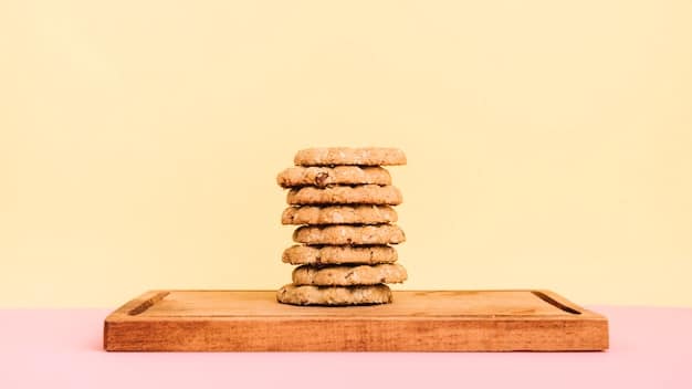 A batch of freshly baked peanut butter cookies on a wooden board. The cookies are golden brown with visible peanut pieces. The background includes a jar of peanut butter and a whisk, suggesting the simple ingredients used.