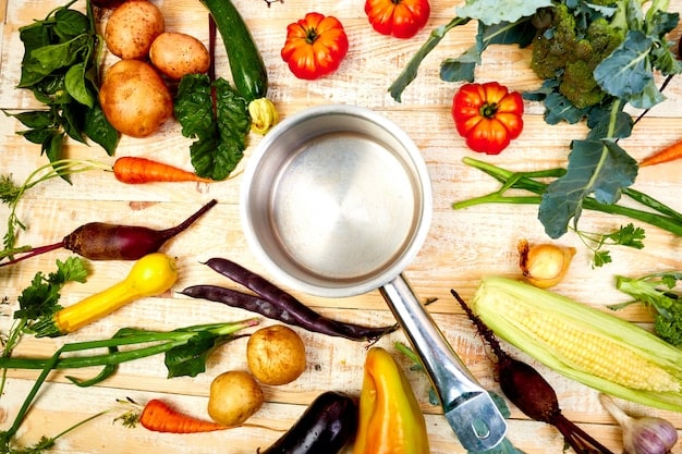 A stainless steel Dutch oven sits on a stove top, surrounded by wooden spoons, measuring cups, and various fresh vegetables like carrots, celery, and onions, ready to be used in a one-pot meal.