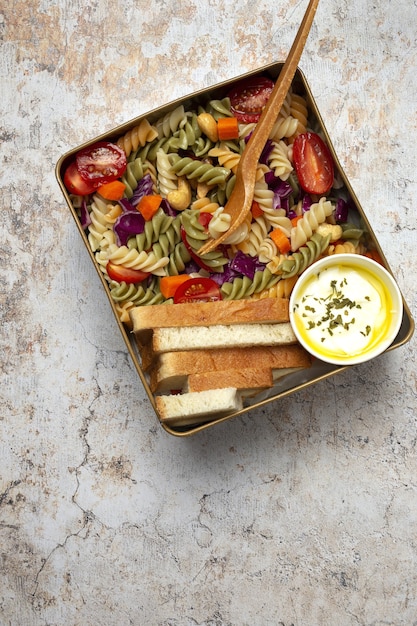 A close-up shot of a perfectly packed lunchbox featuring a colorful pasta salad in a jar, a small container of grapes, and a reusable water bottle. The lunchbox is sitting on a school desk with an open textbook beside it.