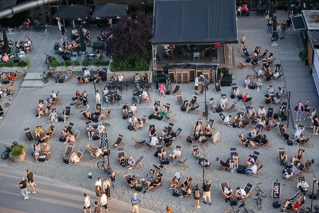 A wide shot of a diverse crowd of people enjoying an outdoor concert in a park. A band is playing on a stage, and families are sitting on blankets, clapping and singing along. Food trucks and vendor booths are set up around the perimeter of the park.