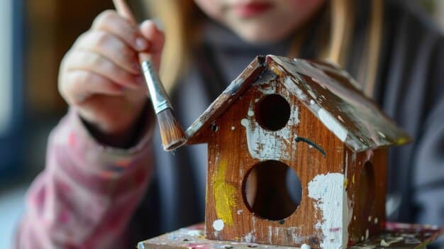 A close-up shot of a child's hands carefully painting a wooden birdhouse. The child is using bright, vibrant colors, and the background shows other art supplies, suggesting a creative and engaging activity.