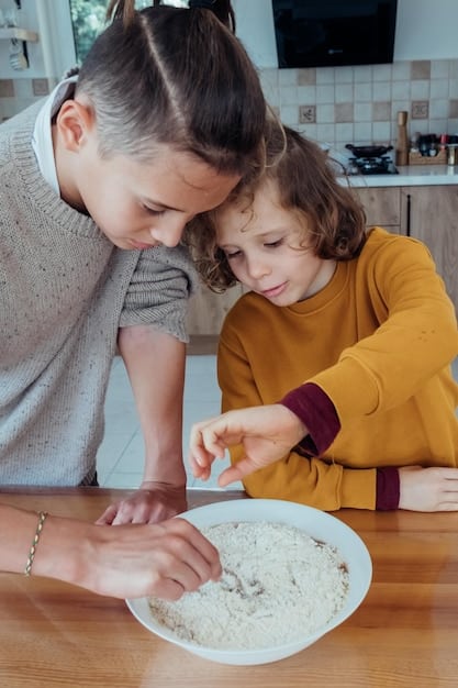 Close-up shot of a child's hands carefully measuring flour into a bowl, with a parent's hand gently guiding them. The background is slightly blurred, focusing the attention on the act of measuring and the connection between parent and child.