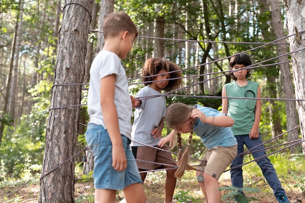 A group of children engaged in imaginative play outdoors, building a fort out of sticks and branches in a wooded area. They are smiling and working together, showcasing teamwork and creativity. The scene is filled with natural light and greenery.