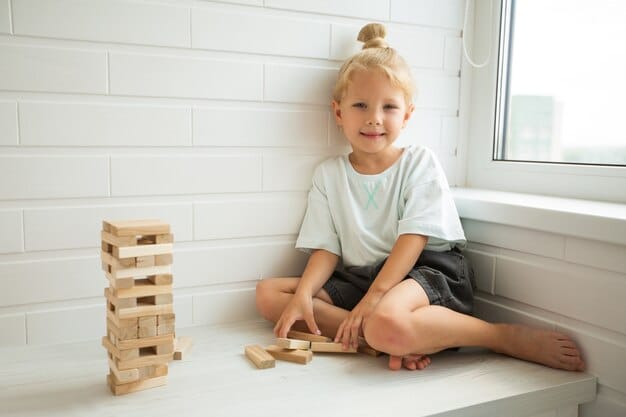 A child smiling brightly while building a tall tower of blocks on a wooden floor. The background is a cozy living room with natural light and soft, inviting furniture.