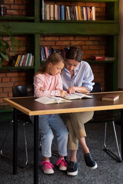 A parent and a teacher sitting at a small table in a classroom, looking at a child's artwork together. They are both smiling and engaged in a positive conversation, with an open and supportive atmosphere. Books and educational materials are visible in the background, enhancing the educational context.