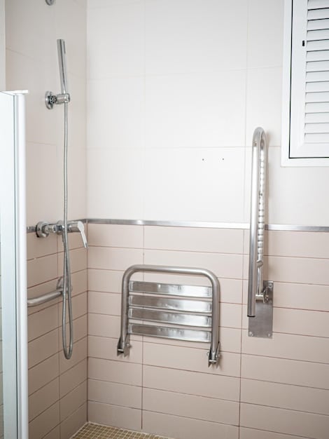 A brightly lit bathroom with grab bars installed near the toilet and in the shower. A non-slip mat is on the floor, and a shower chair is visible inside the shower stall.