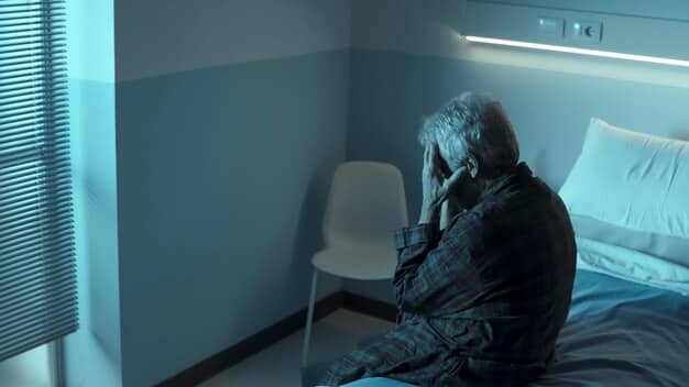 A lone caregiver, head in hands, sitting on the edge of a bed in a dimly lit room, illustrating the isolation and sadness associated with caregiver depression.